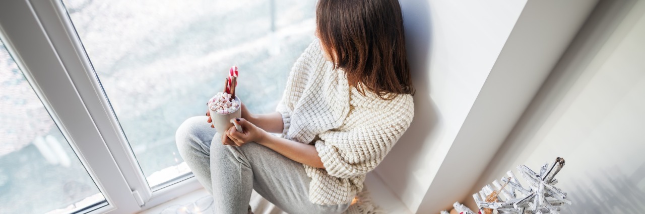 woman dressed in white sitting on a windowsill holding a cup of hot chocolate and looking at the snow