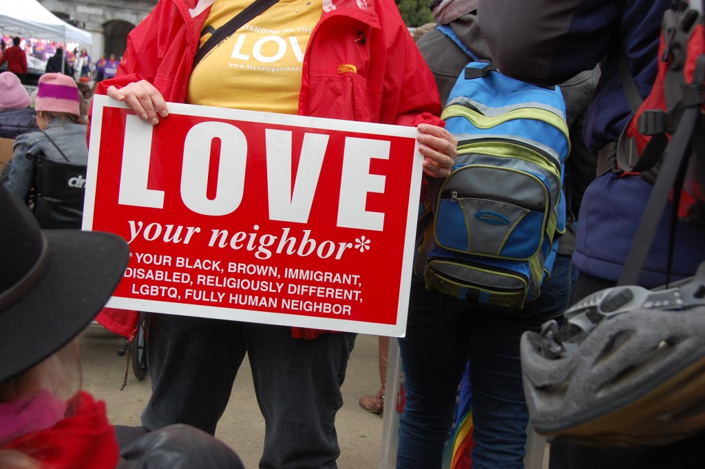 Woman carrying a "love your neighbor" sign listing various groups including disability. 