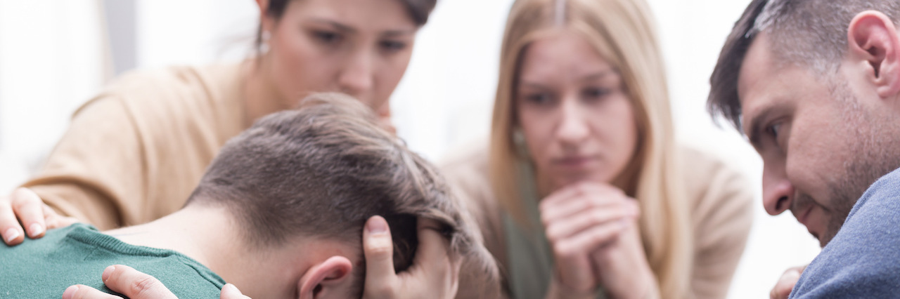 Close-up of a devastated young man holding his head in his hands and a group of friends in a supportive pose around him