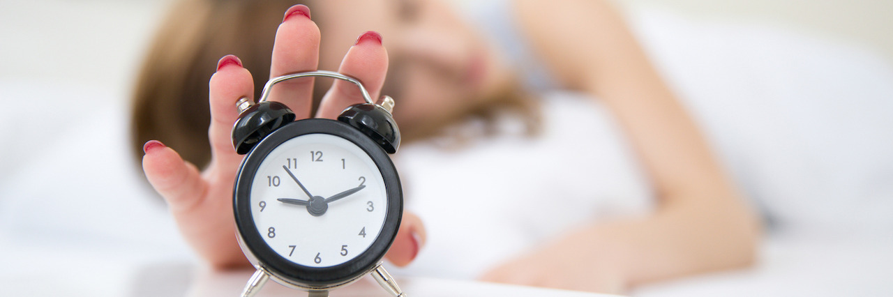 Sleepy woman in bed extending hand to alarm clock. Focus on clock