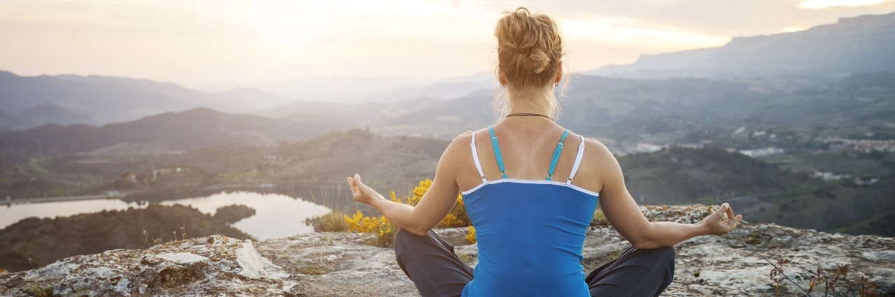 woman sitting on a rock in a meditation pose looking out at mountains and valley