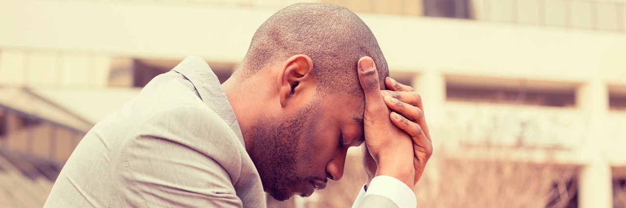 Response to Vicks Study: One in Five Exaggerate Symptoms of Illness side profile young businessman sitting outside corporate office holding head with hands looking down