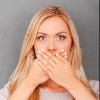 surprised young blond hair woman covering her mouth and staring at camera while standing against grey background