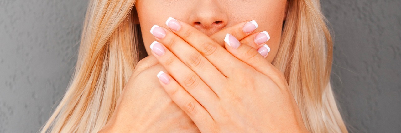 Asperger's Syndrome: Saying Too Much surprised young blond hair woman covering her mouth and staring at camera while standing against grey background
