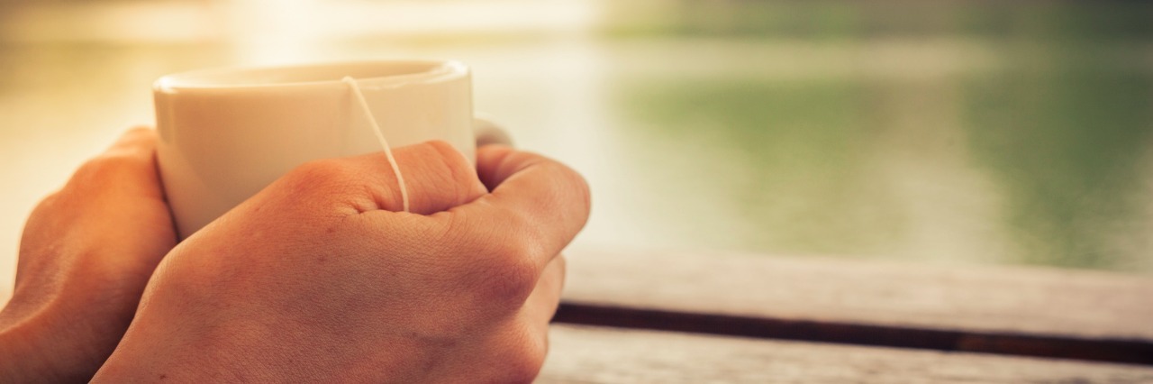 Rheumatoid Arthritis: A Message to Well-Wishers in the New Year closeup on a womans hands holding a cup of tea by a lake in the afternoon
