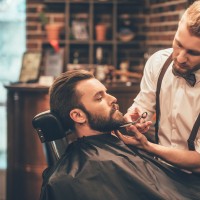 Side view of young bearded man getting beard haircut by hairdresser while sitting in chair at barbershop