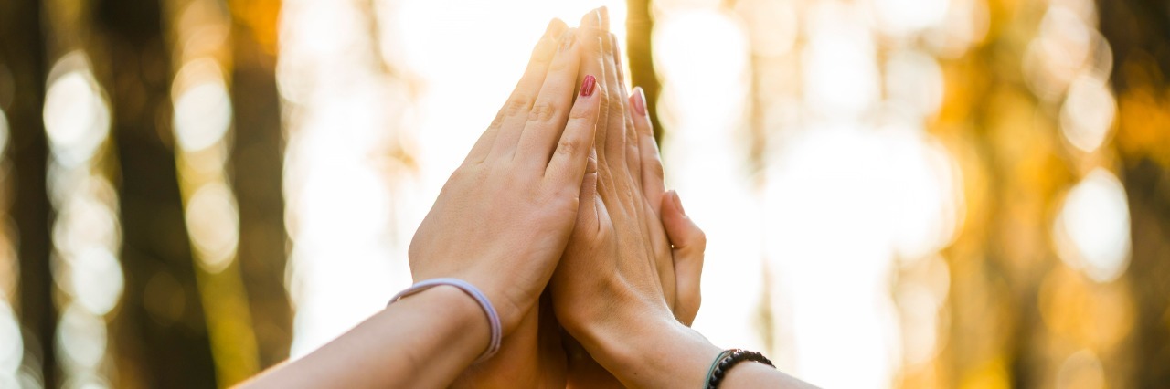 10 Victories Only IBD Patients Understand Closeup view of four people joining their hands together high up in the air outside in a forested area.