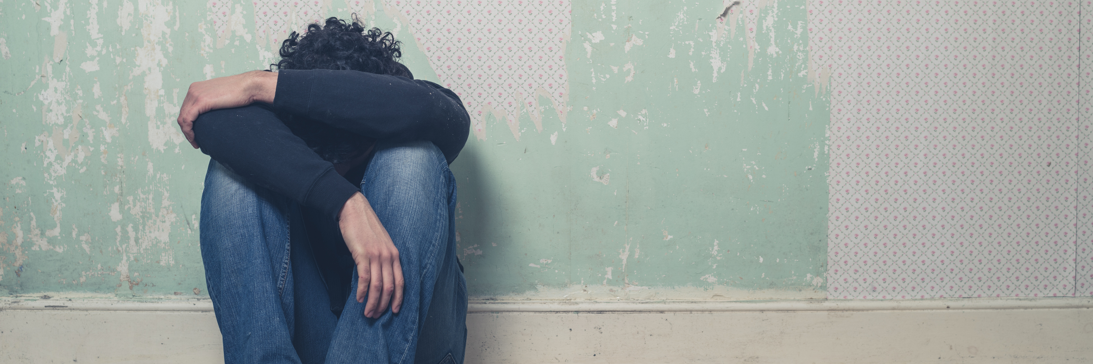 depressed young man sitting on floor in empty room with peeling wallpaper