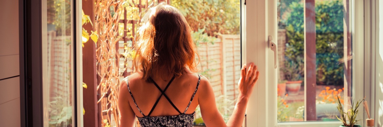 5 Truths to Remember If You're Feeling Guilty About Your Illness A young woman is standing in the doorway of her kitchen and is looking at the garden at sunrise