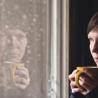 Lonesome woman drinking cup of coffee by the window of her living room