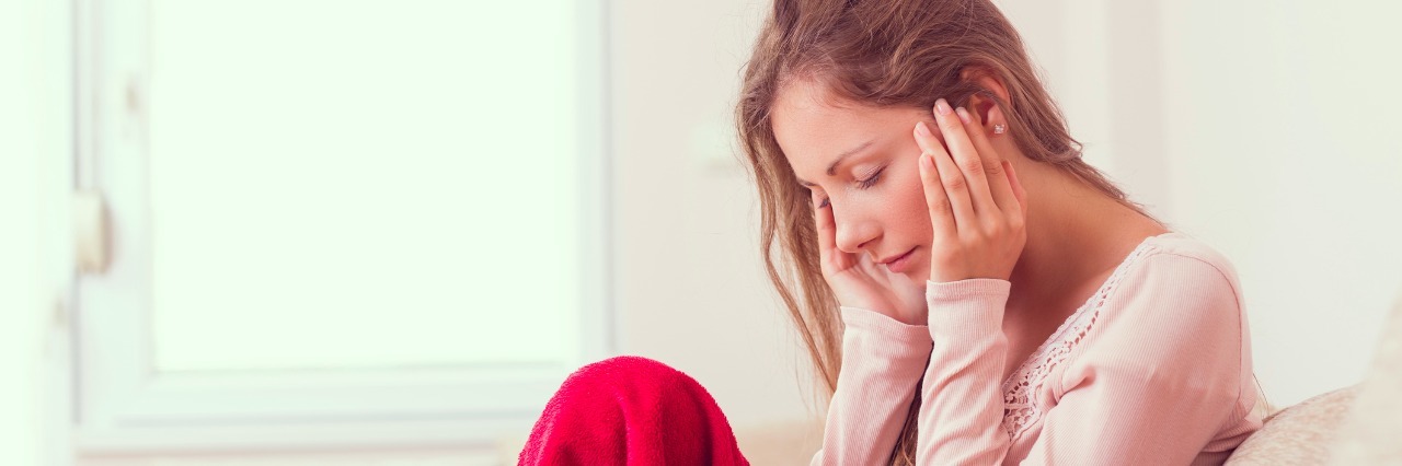 What It's Like Being Home Sick With Hypothyroidism Young woman sitting on a couch, holding her head, having a strong headache. Lens flare effect on the window