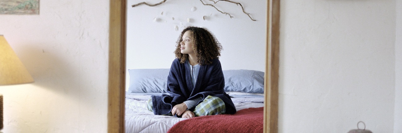 How to Be Independent With Chronic Pain and Fatigue young woman sitting on her bed and looking away, her reflection is seen in a mirror