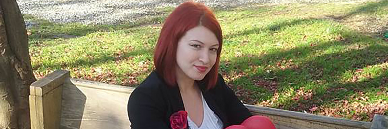 Emily O'Malley sitting on a bench. She has red hair and is wearing a white shirt, black jacket, bright red leggings and black boots.