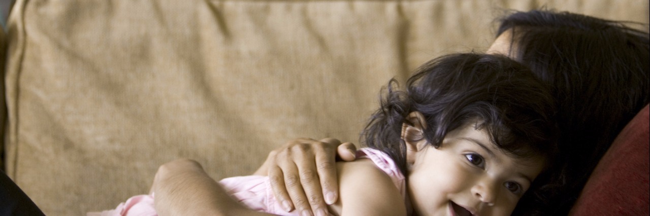 How to Be a Mom With Chronic Fatigue Syndrome lifestyle shot of a mother as she lays on the couch and lovingly hugs her toddler daughter