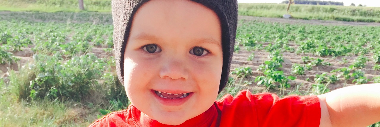 Little boy wearing a bright orange shirt that reads, "Alaska" and a batman beanie-hat on his head