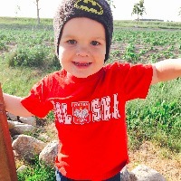 Little boy wearing a bright orange shirt that reads, "Alaska" and a batman beanie-hat on his head