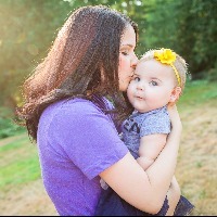 Mom holding baby daughter, kissing her cheek in front of a tree