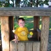 Boy wearing yellow shirt and holding a sippy cup with a matching yellow top playing inside a wooden play structure.