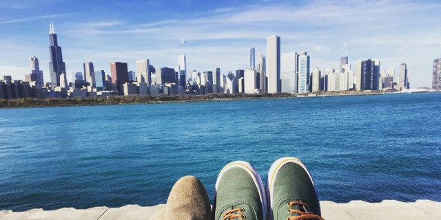 A man and woman's feet, facing a body of water and a city skyscraper.