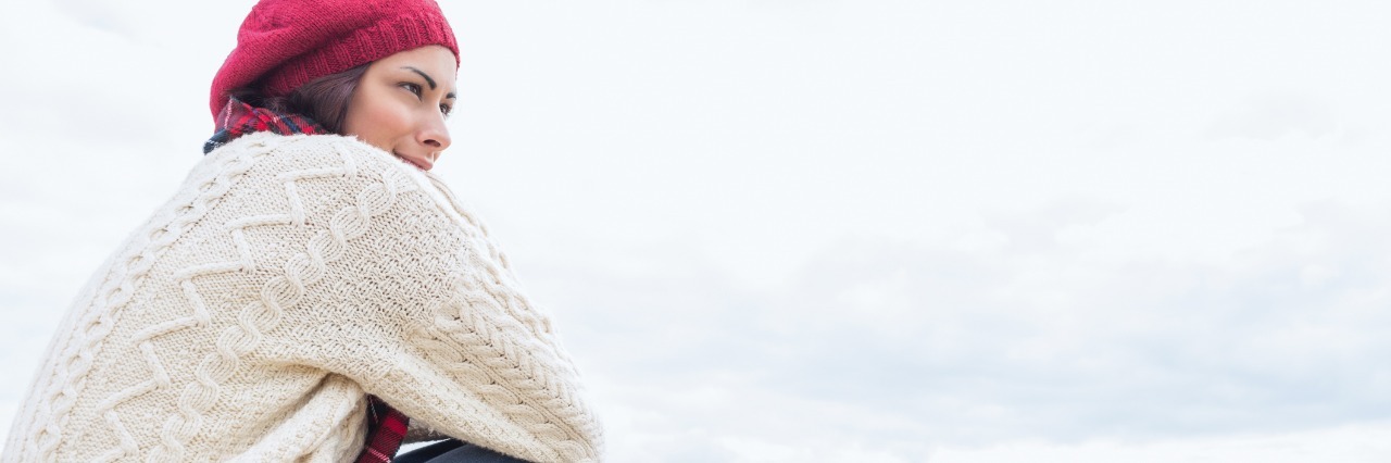 Letter to Person Struggling With Chronic Illness woman in sweater and jeans sitting on the beach and hugging her knees