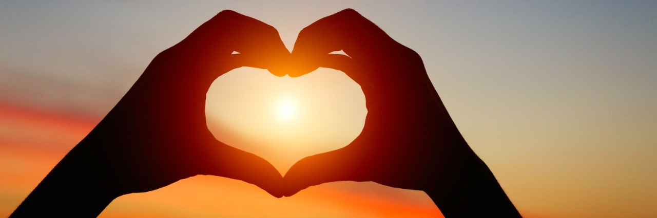 Defining Self-Love in My Life With Physical and Mental Illness Two hands making a heart sign against a sunset sky