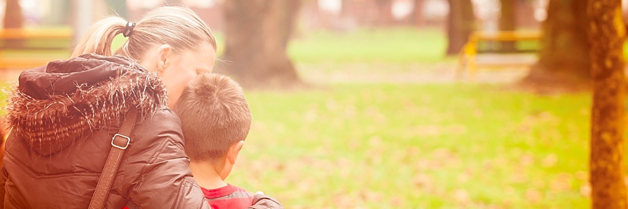 Mother and son walking at the park. Mother is embracing her son with head leaning against his.