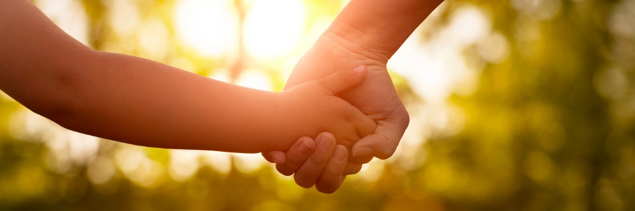 Parent and child holding hands outdoors with trees and sunlight in the background