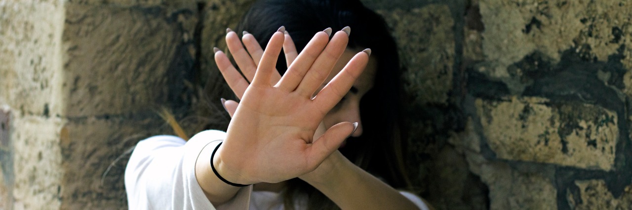 Myasthenia Gravis: Fearing Others Won't Believe My Illness woman standing in front of a stone wall and holding her hands up to block her face