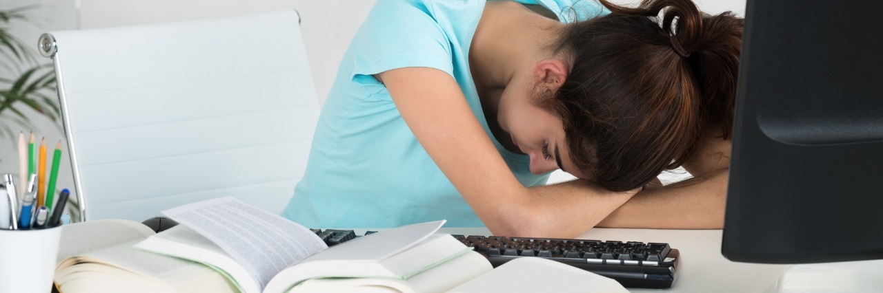 Relief of Knowing My Exhaustion is Due to POTS woman resting her head and arms on her desk next to a computer and books