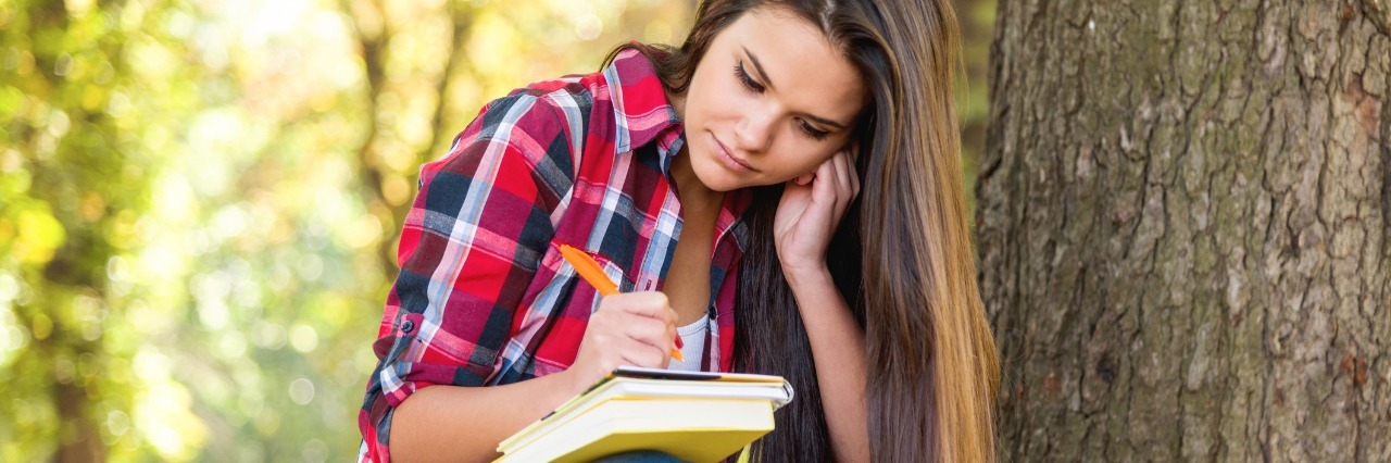 college student sitting on a bench outside next to a tree with books and a journal