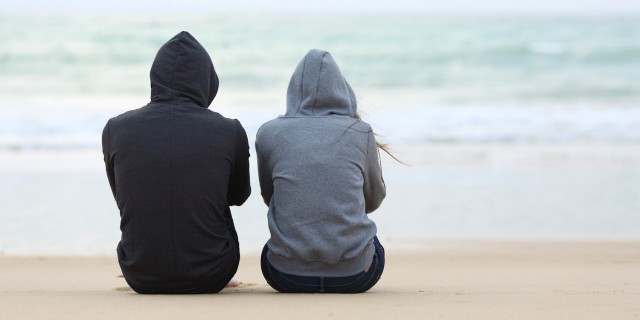 Two people wearing hoodies, sitting on sand on beach facing ocean