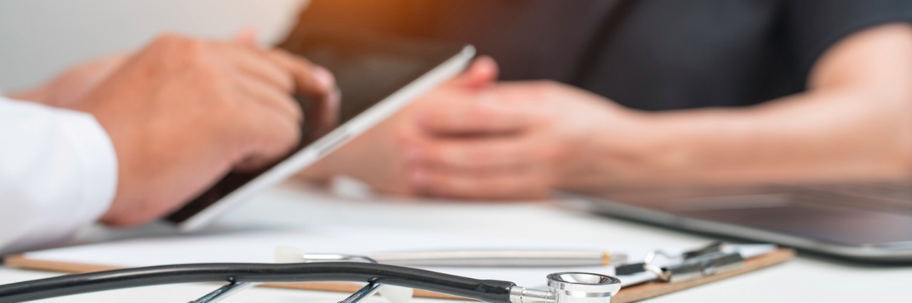 To the Geneticist Who Knows a Condition but Not My Son Close-up of doctor holding tablet, sitting at desk across from patient