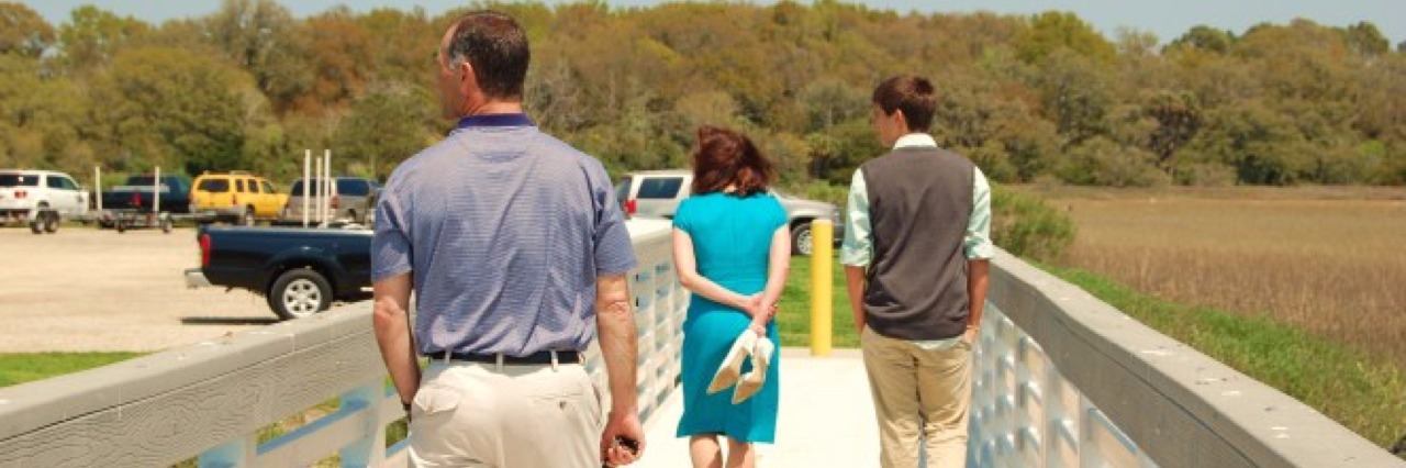 Grieving 18 Months After Suicide Loss Three people, two men one woman in a bright blue dress, walking on a footbridge. They are facing away from the camera.