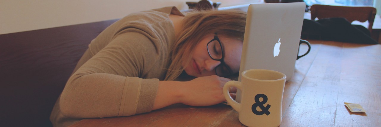 Why I Blog About Having Chronic Illnesses A woman sits at a table, her head resting on her laptop, coffee mug nearby.