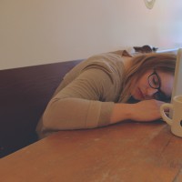 A woman sits at a table, her head resting on her laptop, coffee mug nearby.