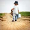 Back of little boy walking on a path, he is wearing a hat, and holding a teddy bear and a tiny suitcase