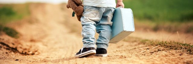 Back of little boy walking on a path, he is wearing a hat, and holding a teddy bear and a tiny suitcase