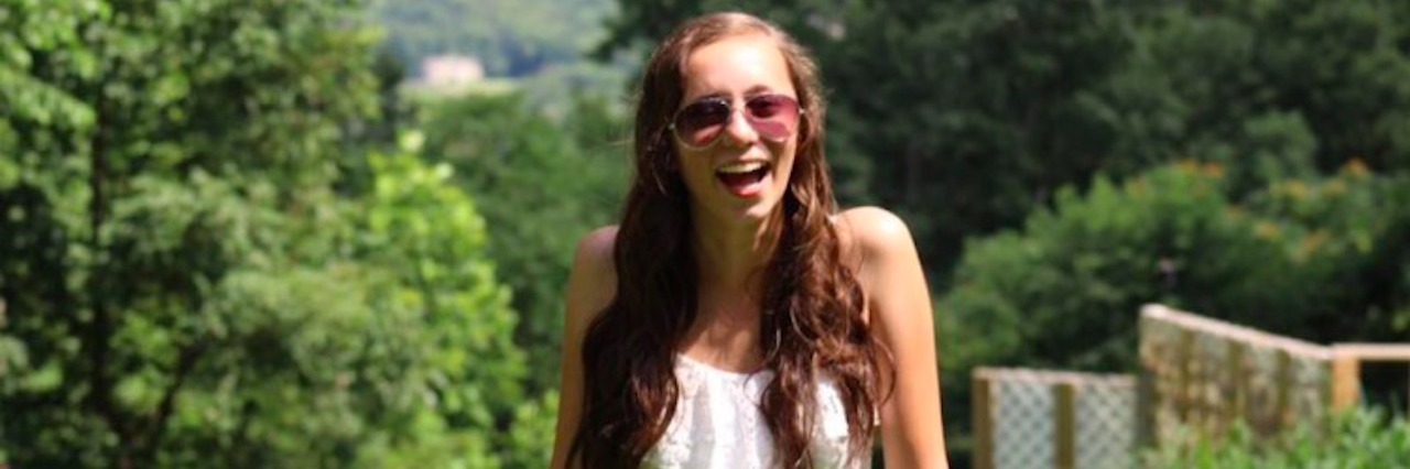 A woman in sunglasses and a white sun dress is smiling, with nature behind her.
