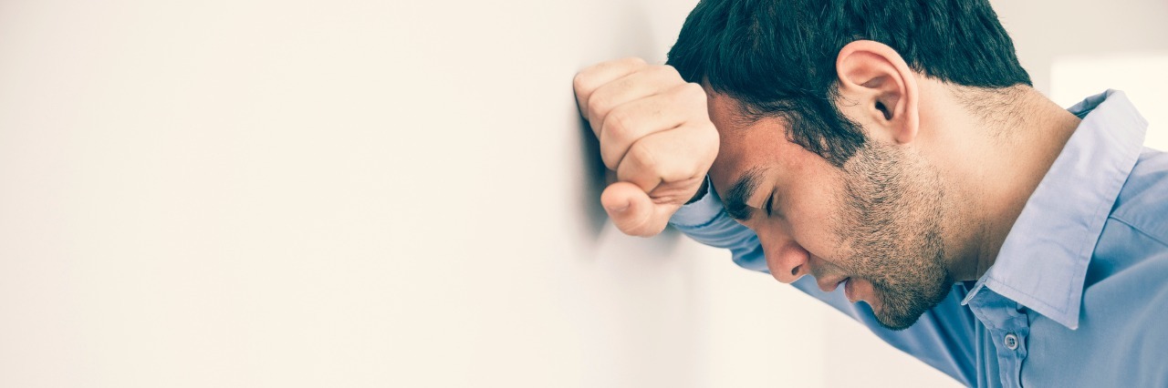 Letting Go of the Shame I Feel Sharing About My Spouse as a Widower A man with a bear and dark hair, wearing a blue shirt, is leaning his head against a wall and with his head against his arm.