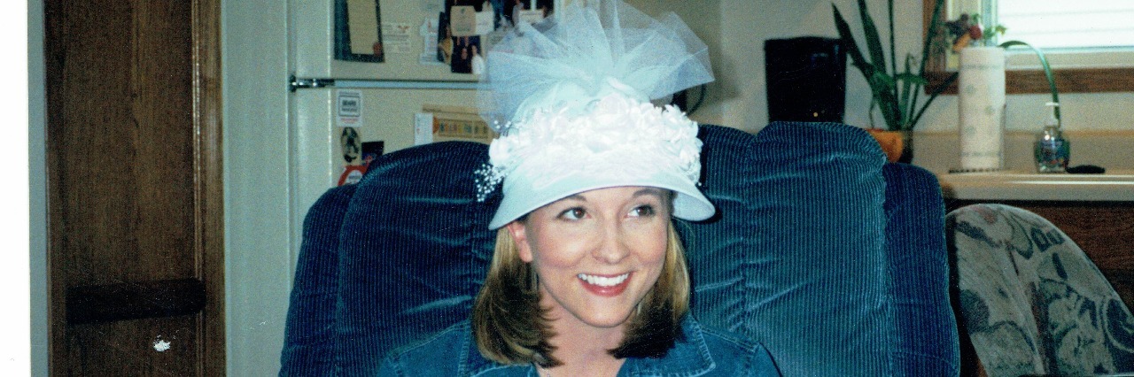Woman sitting on a blue chair and wearing a denim dress and a big white hat with a big bow on top.
