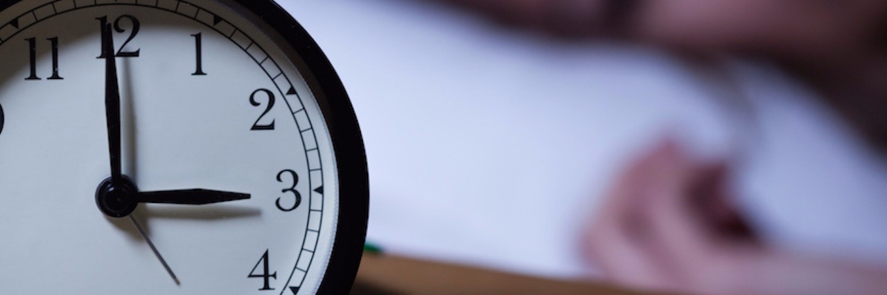 Earthquake Anxiety and Grief in the Middle of the Night Alarm clock on bedside table set to 3 and woman in bed in the background, looking toward the clock