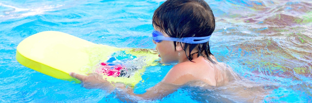 Boy learning to swim in swimming pool, wearing goggles and holding a kickboard