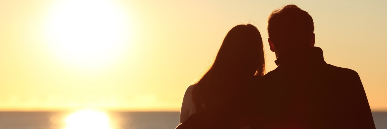 silhouette of woman and man sitting on a bench in front of the ocean watching the sunset