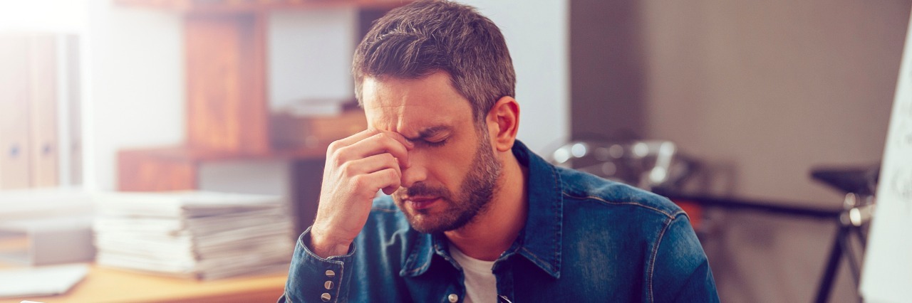 young man massaging his nose and keeping eyes closed while sitting at his working place in office