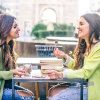 Two women friends laughing while sitting in a bar outdoors - Students having pause and drinking cappuccino - Best friends talking and having fun