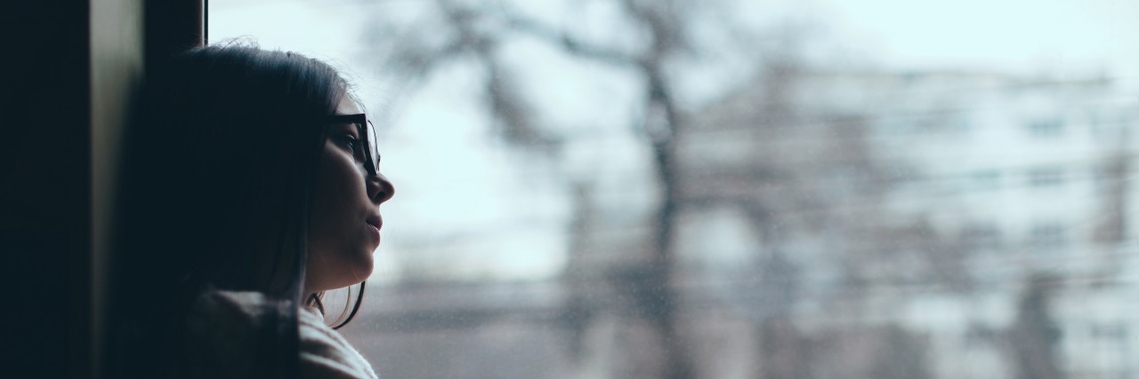 Woman with long dark hair and thick glasses looking out a window.