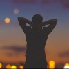 Silhouette of a man with hands folded behind head, with evening sky and city lights in the background
