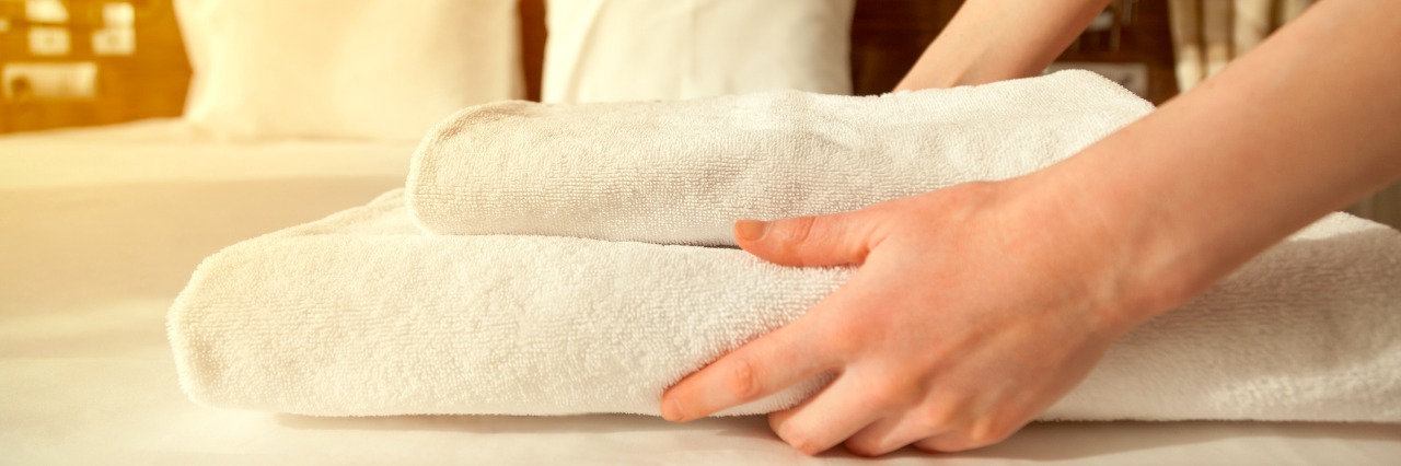 Close-up of hands putting stack of fresh white bath towels on the bed sheet. Room service maid cleaning hotel room. Lens flair in sunlight