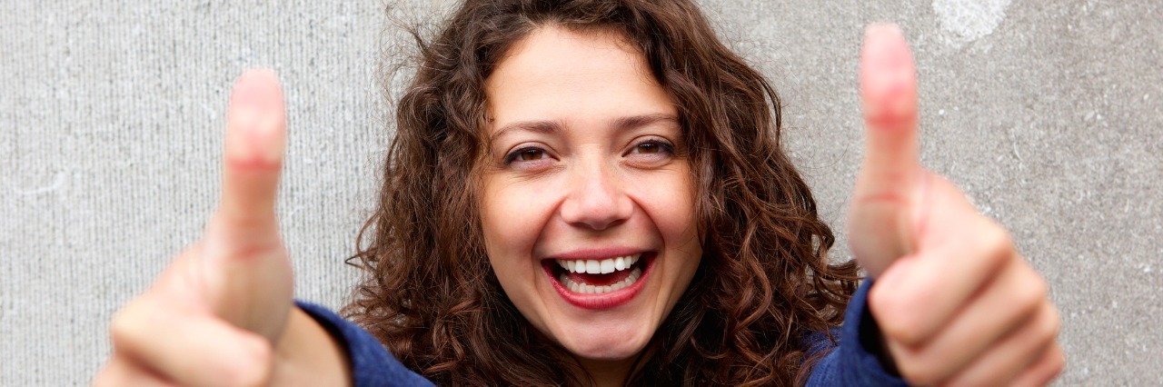 6 Reasons You're Awesome If You Have Chronic Fatigue Syndrome Portrait of smiling young woman giving thumbs ups with both hands against gray wall