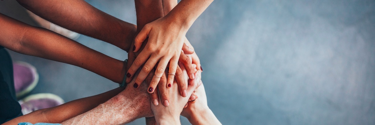 How My Family Can Help Me Face My Chronic Illness Close up top view of young people putting their hands together. Friends with stack of hands showing unity.